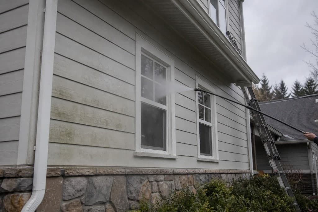 Technician on a ladder washing house siding above mortared stone wainscot and foundation plantings.